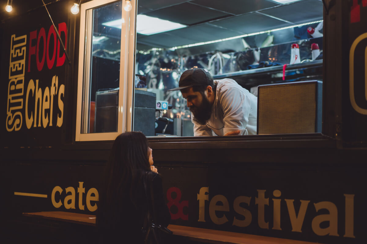 Bearded street food chef leaning out of a 'Street Food Chef's' catering truck window to serve a customer at a festival - inspiration for naming a food truck business